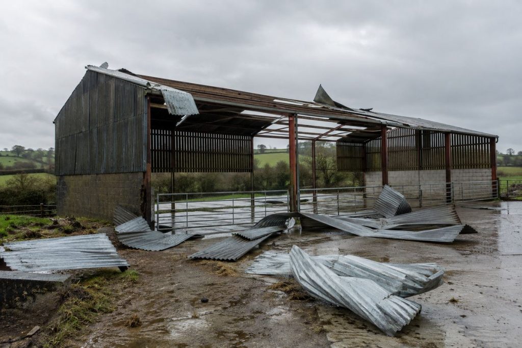 Storm-damaged farm shed with roof sheeting torn off by high winds and twisted corrugated sheets across the yard